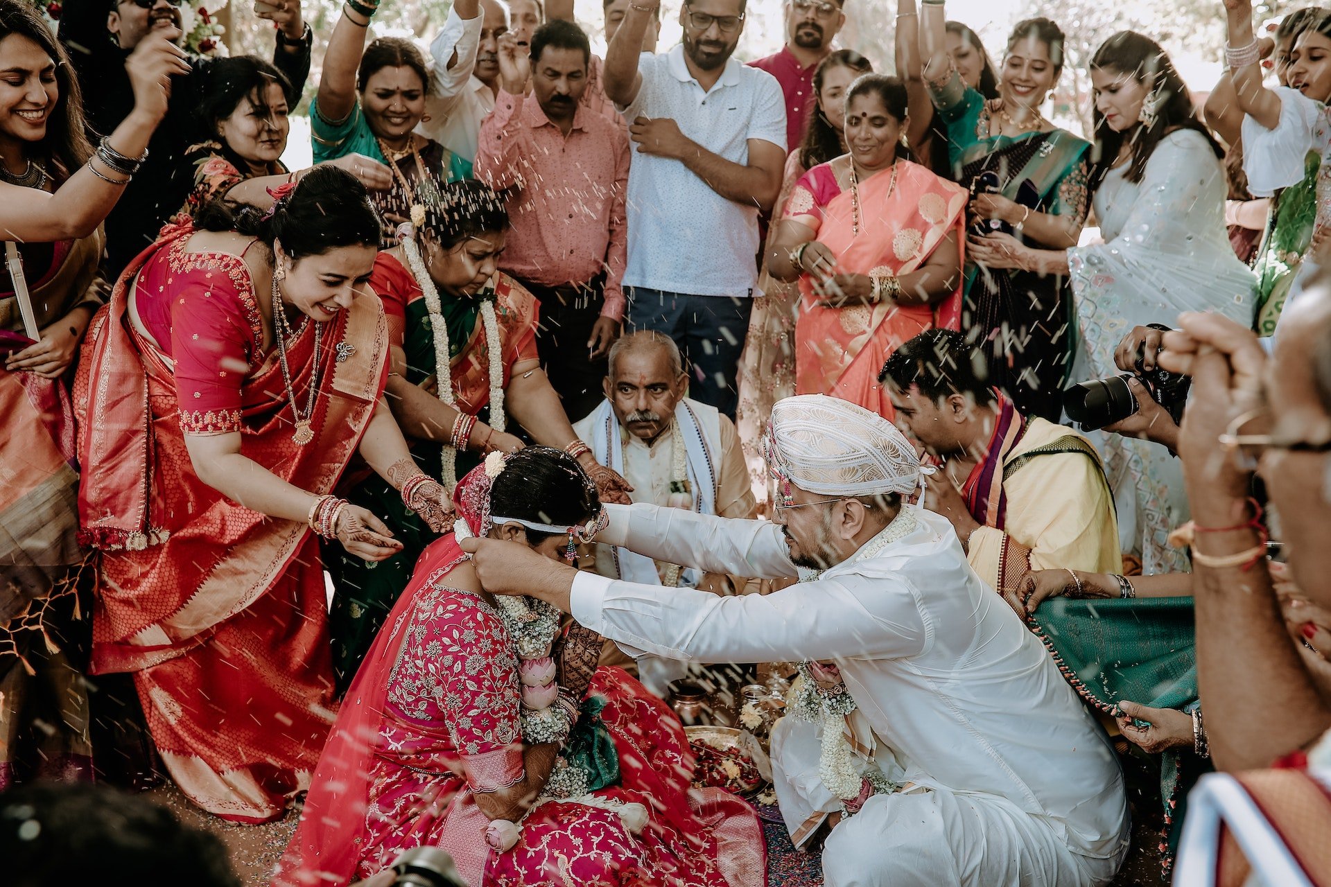 Depicts a couple at an Indian wedding. They are exchanging flower garlands surrounded by family throwing rice.